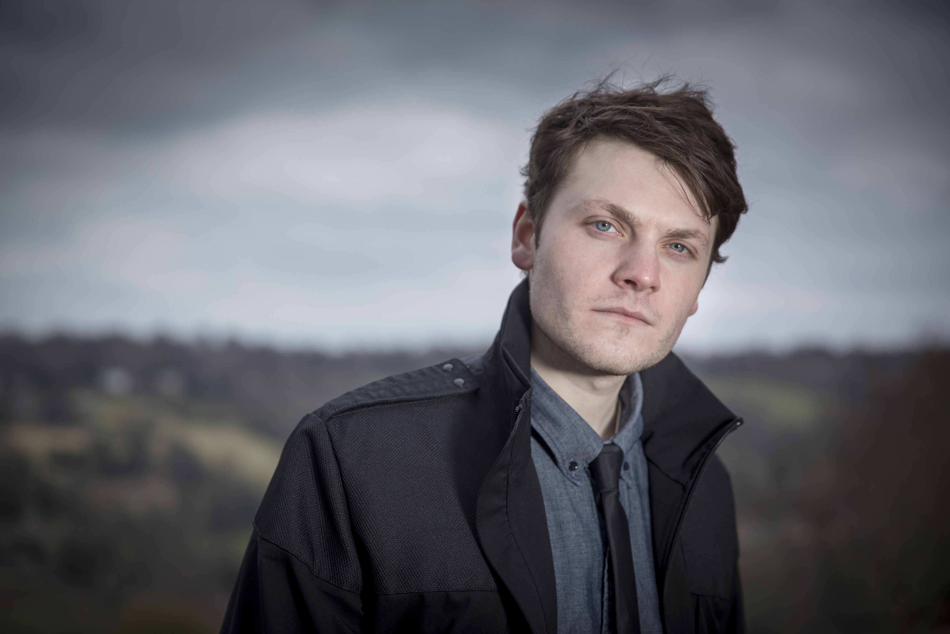 White man with dark hair, shirt and tie and black jacket, posing against a backdrop of a hillside and sky