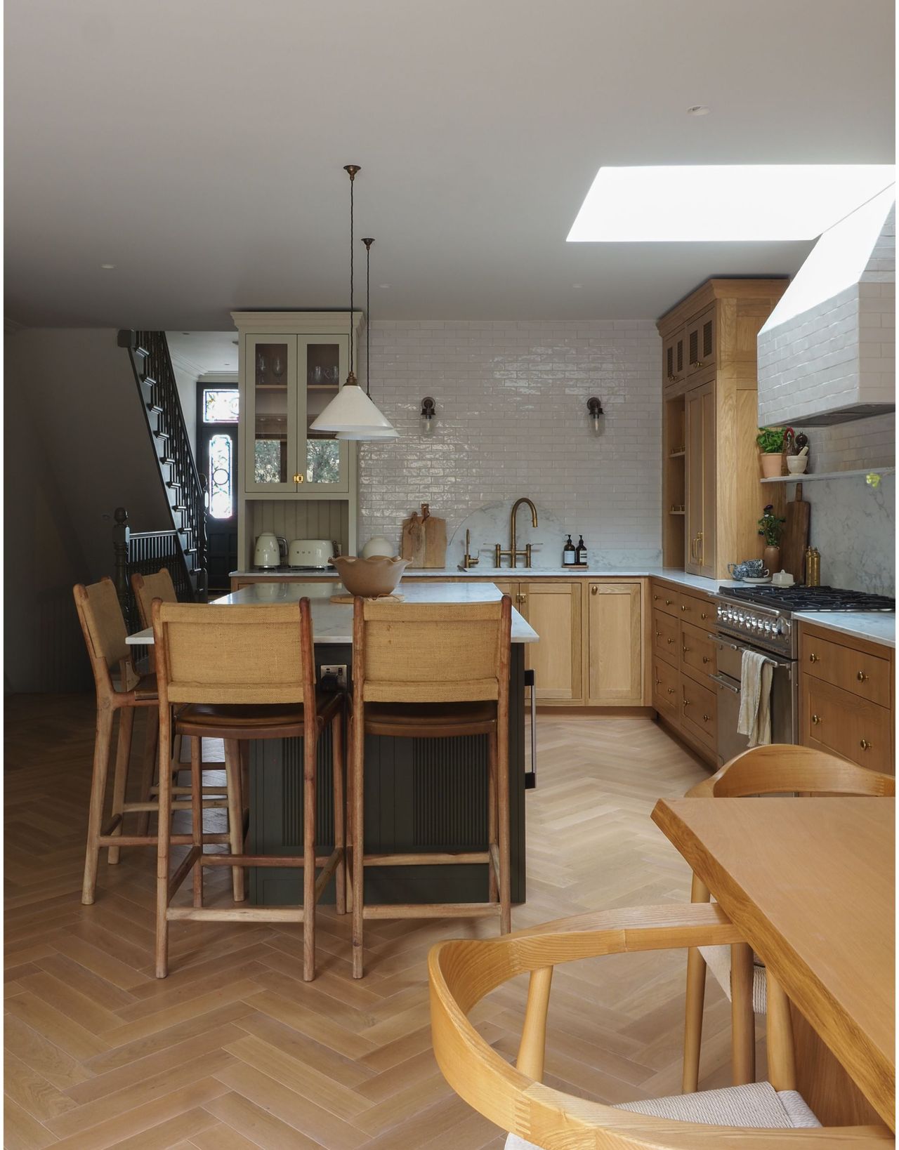 A modern kitchen with kitchen island seating with wooden bar stools and two pendant lights hanging over the island in a wood and white themed space with stairs and an entryway in the background