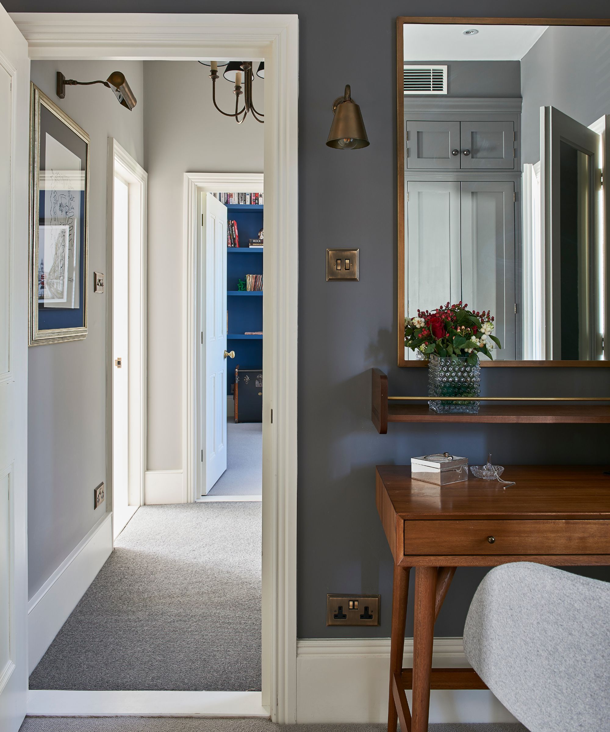A grey room with white baseboards, white doorway trim, a large wooden mirror, wooden table, and gold lightswitch and gold light above. To the left is an open door to a hallway seen beyond.