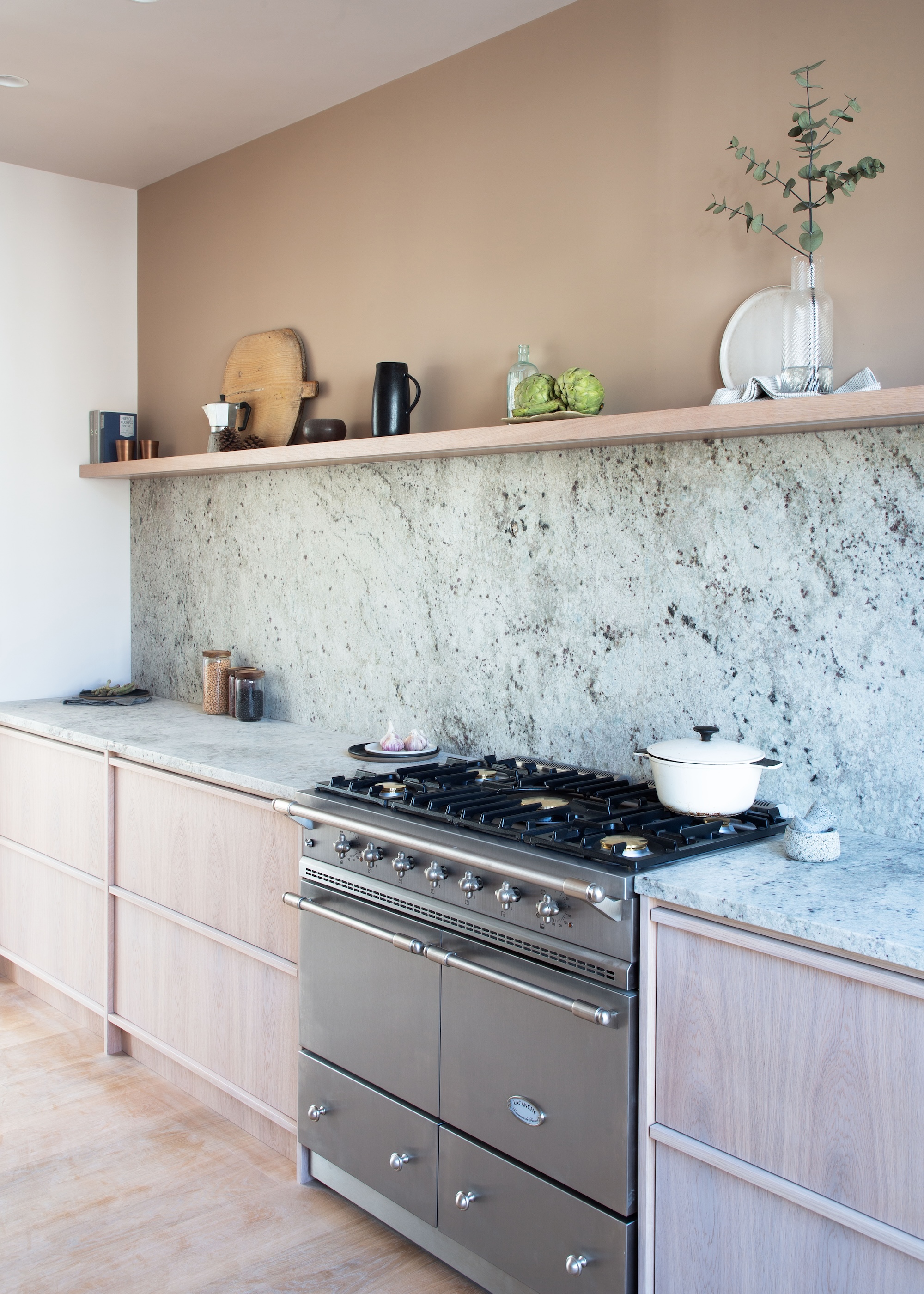 A pastel colored kitchen with a view of a gas stove, on top of the stove is a white pot and beside it are a range of kitchen accessories. Above the stove is an open shelf with other kitchen essentials and accessories.
