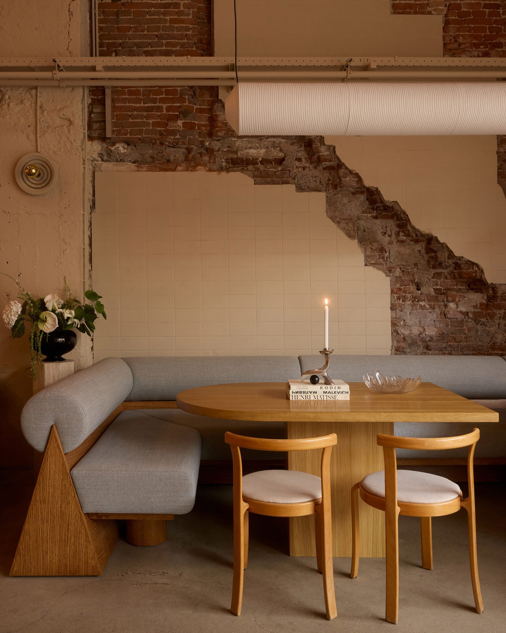 Interior of a restaurant in an industrial site, with gray fabric and pale wood banquettes, wooden furniture, a lit candle in a sculptural silver candleholder, and fresh lily flowers.