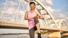 Woman in sportswear raises one knee to meet her hands. She is outside with a bridge behind her