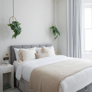 A neutral guest bedroom with double bed, beige throw and cushion decor, white bedside table, light grey curtain window treatment, bay windows and faux foliage adorning brass pendant light fixtures