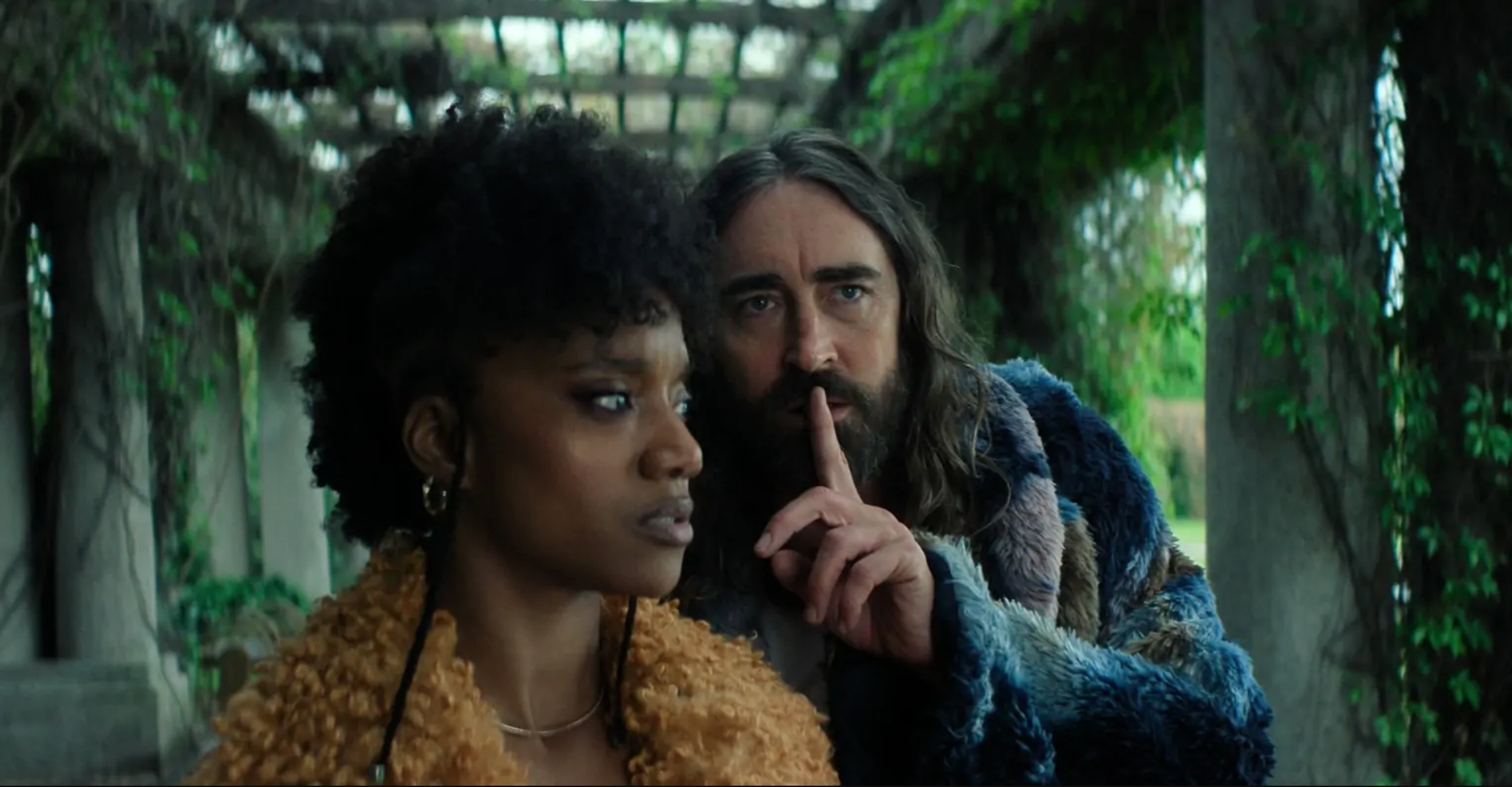 a man makes a silencing gesture to a woman in a garden