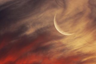 A large crescent moon hangs in the top right, wistfully obscured by the flow of passing clouds, illuminated orange from the rays of an unseen setting sun.