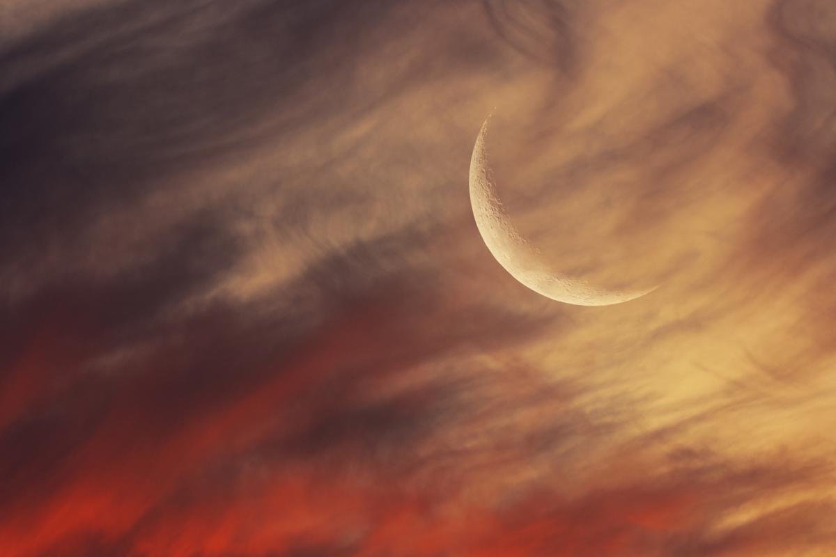 A large crescent moon hangs in the top right, wistfully obscured by the flow of passing clouds, illuminated orange from the rays of an unseen setting sun.