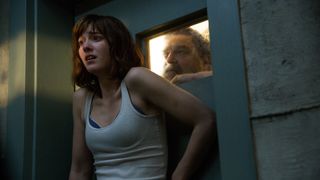 A woman looks scared while a man observes her through a small window in a bunker