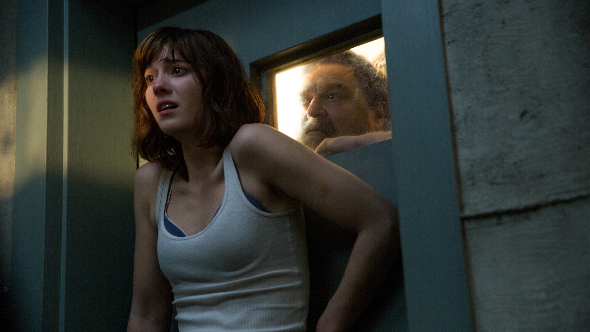 A woman looks scared while a man observes her through a small window in a bunker