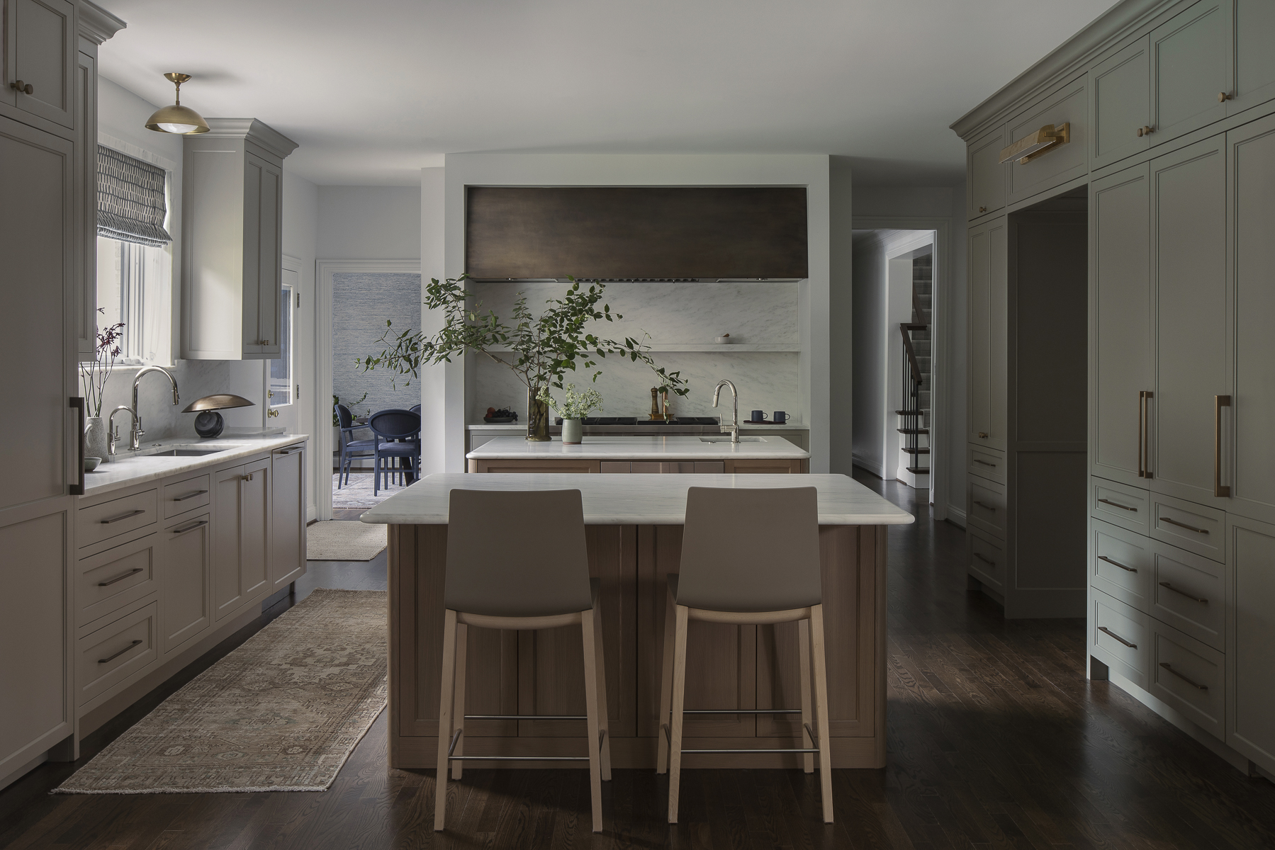 traditional style kitchen with light olive green cabinetry on one side, and cream on the other. the island bench is timber with white counters and two gray bar stools, and the backsplash is white stone with a brass range hood