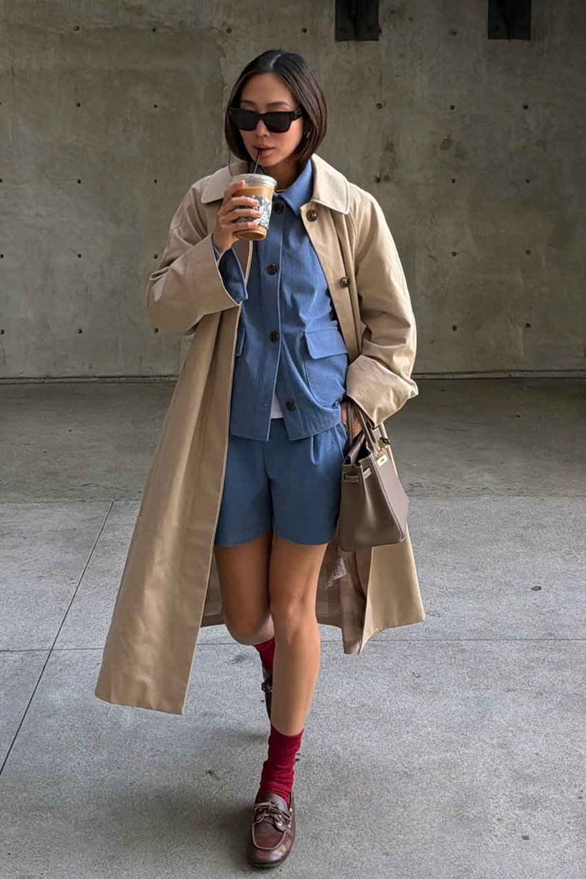 a woman wearing a trench coat, blue button-down, matching shorts, red socks, and brown loafers
