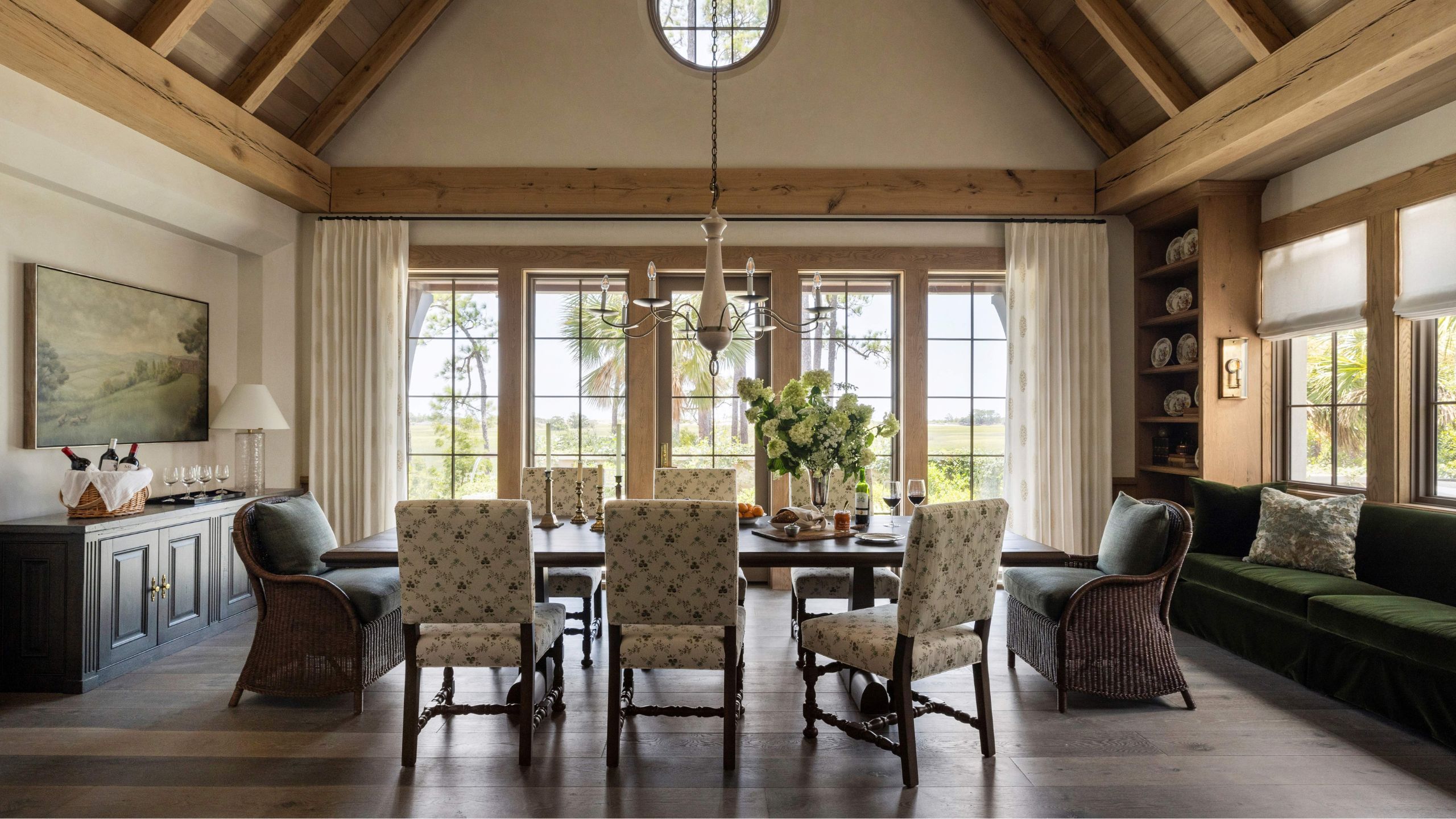 a vaulted pitched roof dining room with glass windows and doors along one, wooden beams, dark wooden floor, a green velvet couch on the right side, and a mix of floral and wicker chairs at the table