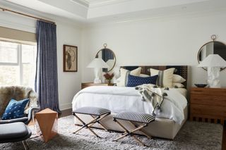 Beige bed covered in white, beige and dark blue bedding and cushions, two striped upholstered stools at the end of the bed, mottled blue curtains and orange geometric side table