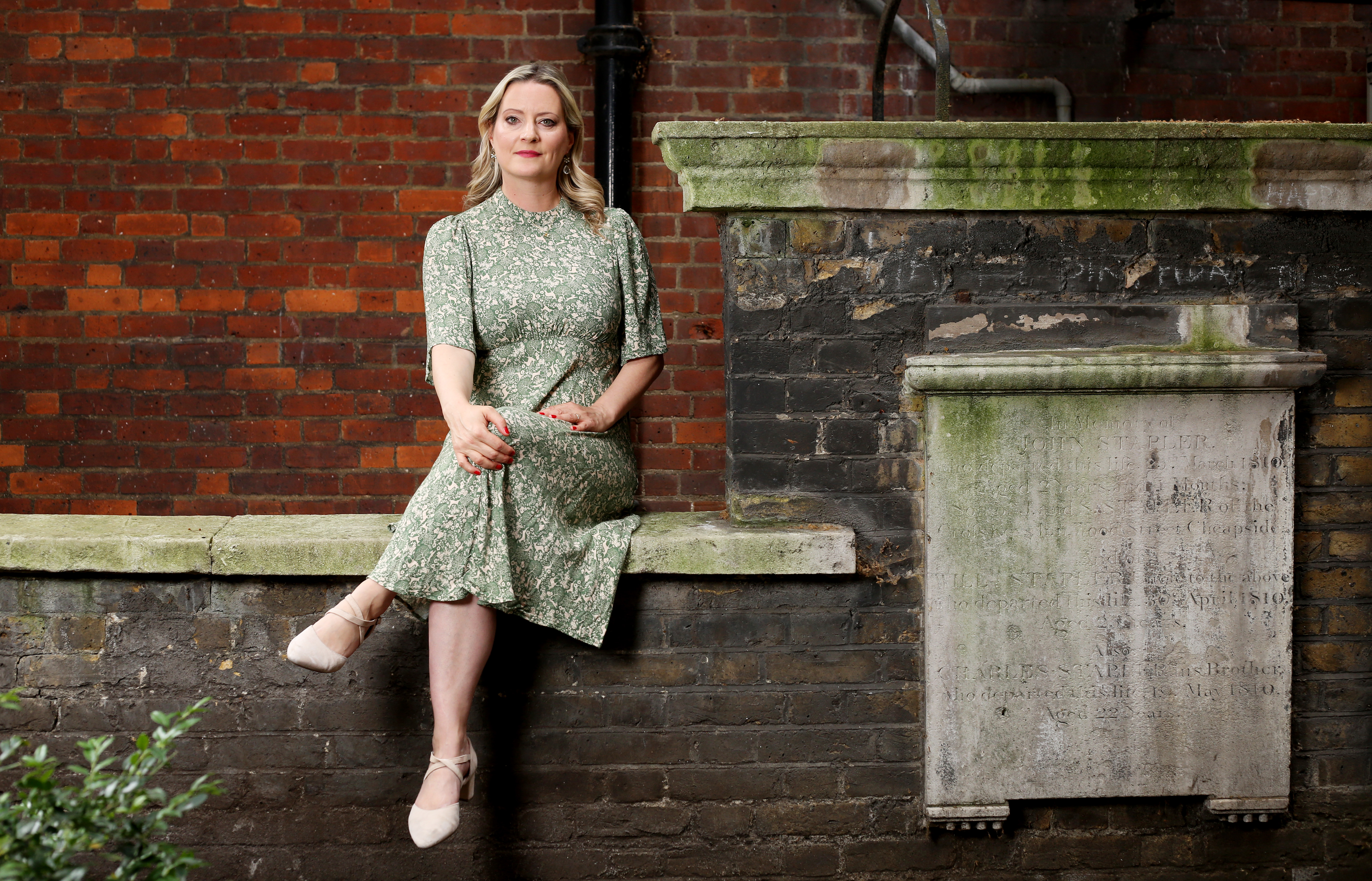 Victoria Shepherd sitting on a dirty brick wall in a green, floral-pattern dress and nude pumps