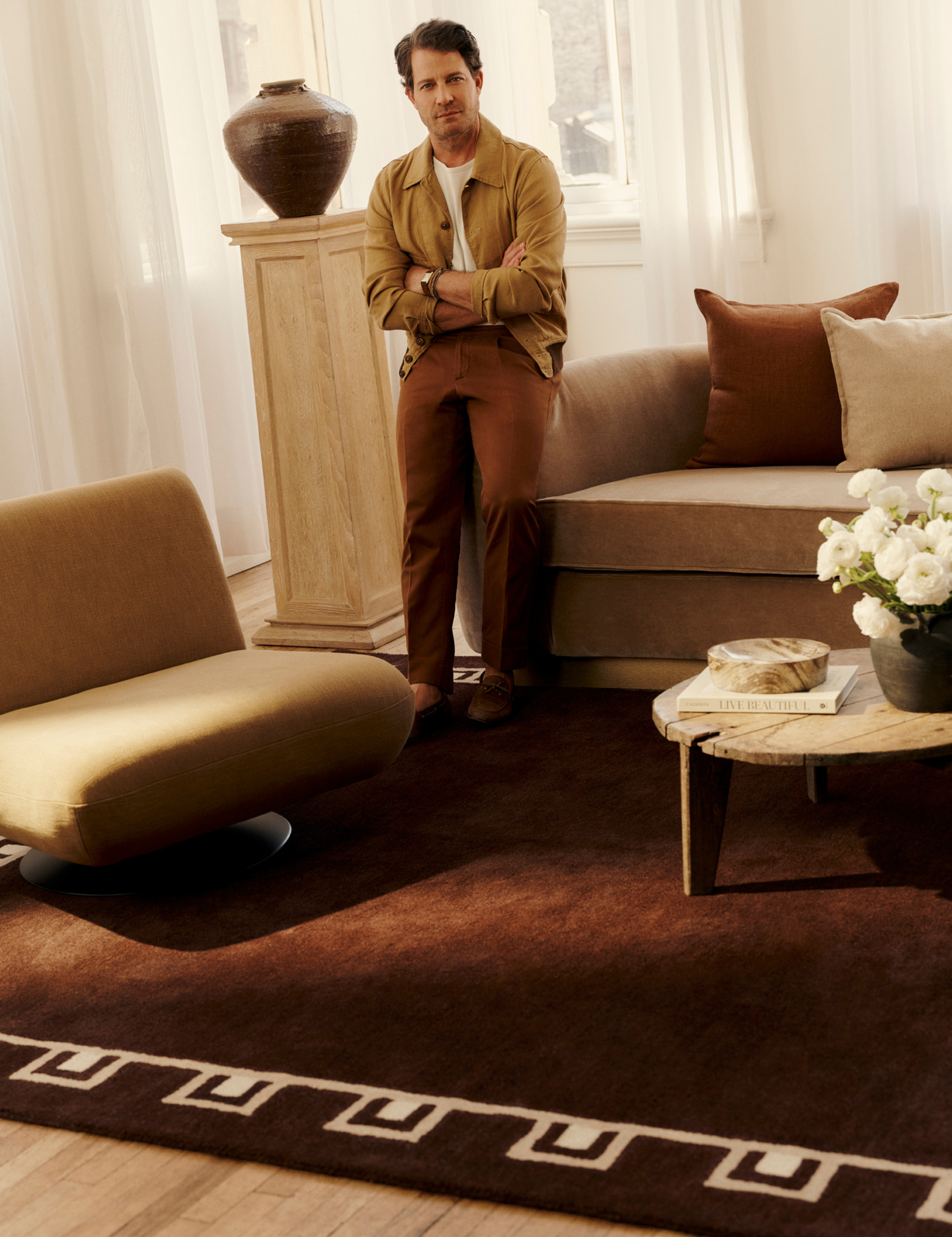 Designer Nate Berkus poses in a warm NYC living room with a chocolate-brown-and-ivory bordered rug from the second release of his Rugs USA collaboration styled underfoot