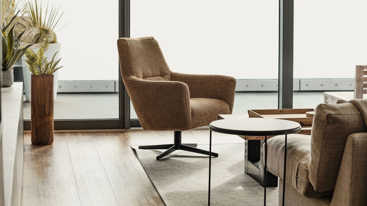 A mid-century modern style arm chair on casters on the corner of a thin area rug, on top of a light wooden floor. The back corner of a couch visible in the foreground, with a small round metal table beside it. large floor-to-ceiling windows in the background. 