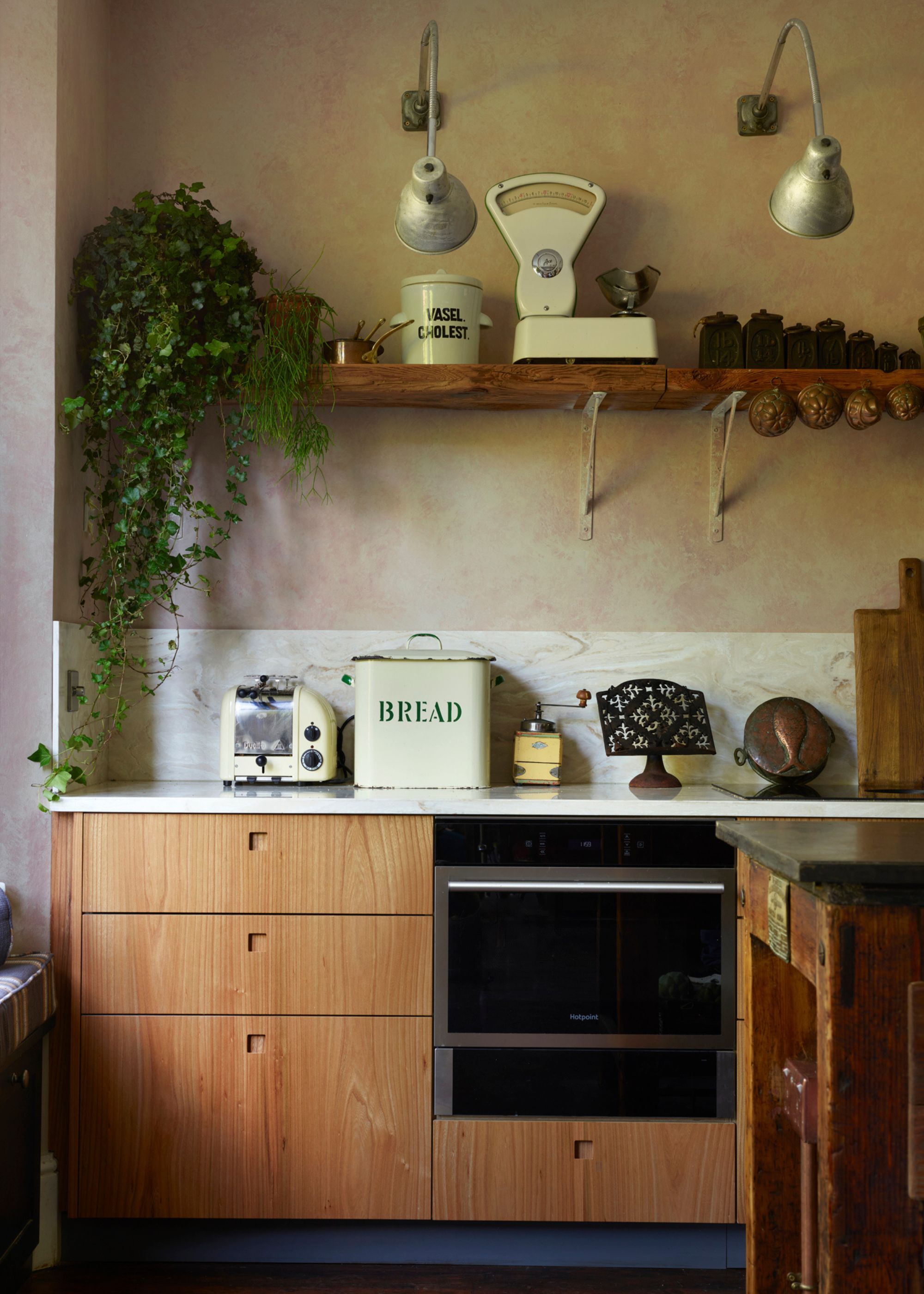 A light oak kitchen with lower cabinets and open shelving above. On the open shelf is a potted plant hanging down with a weighing scale as well as other kitchen essentials. There is also a view of the kitchen oven and stove as well as kitchen counter.