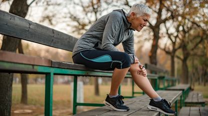 Older lady sits on a bench as she holds her leg in pain