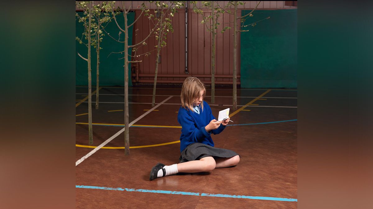 Girl sat on school gym floor playing video game device.