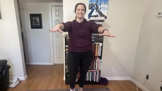 Jennifer Rizzuto performs a walking workout at home. She is dressed in leggings, a hooded jumper and sneakers. She is walking on the spot and smiling, looking at the camera, with her arms held straight in front of her. Behind her we see pictures on the walls and a bookcase.