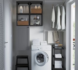 A laundry room with white washing machine with white towels on top, black open shelving with baskets, area to hang shirts, black console table with towels and glass/bamboo containers and black wooden stool