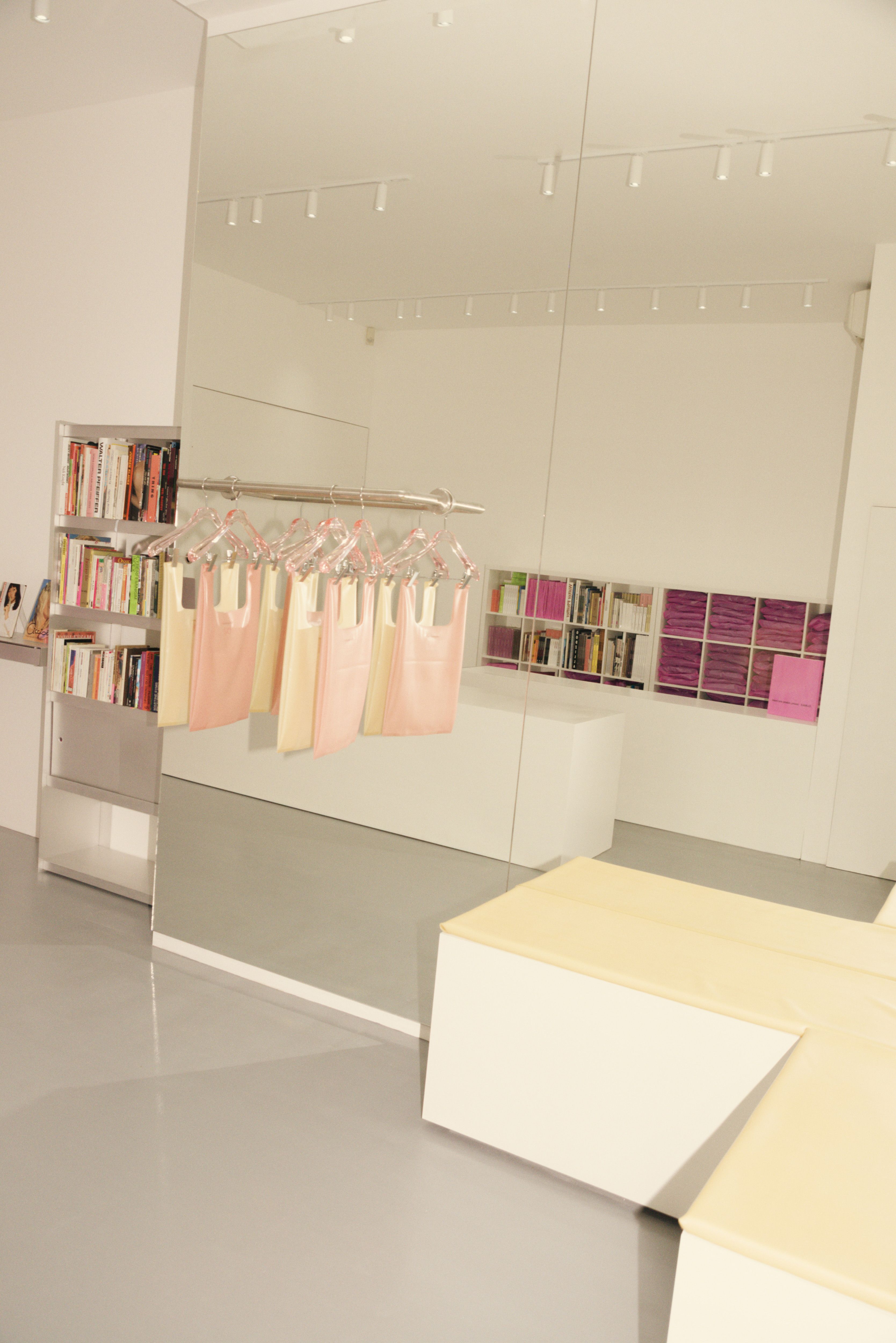 The interiors of a sleekly designed, brightly lit books and merchandise shop features white-washed walls, chrome shelves stakced with colorful tomes, and a pink-hued, see-through door.