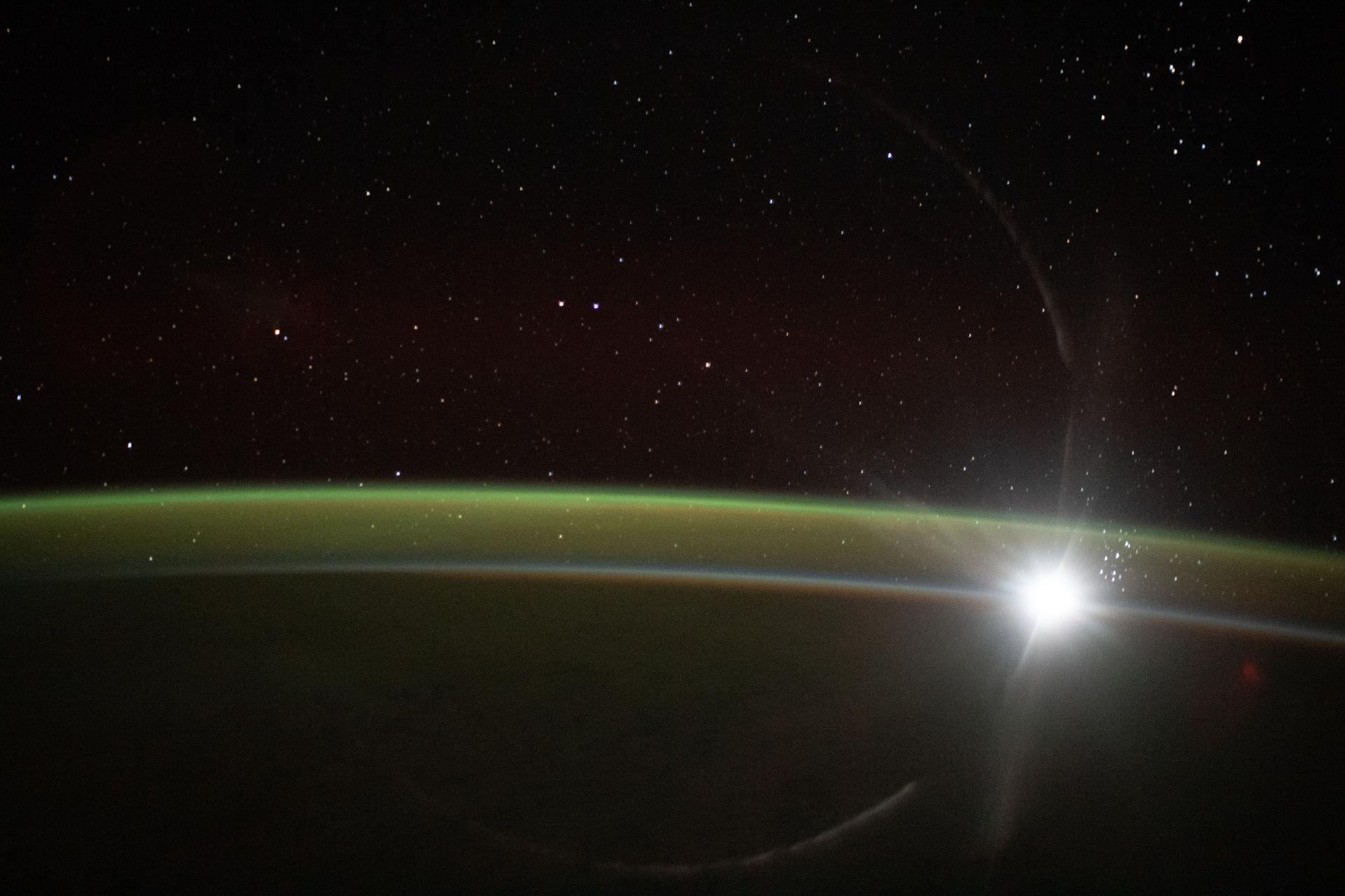 the curve of the earth, shaded in black, is blanketed by a thin green line of atmosphere. the sun peaks from the edge of the planet's curve. stars glimmer above.