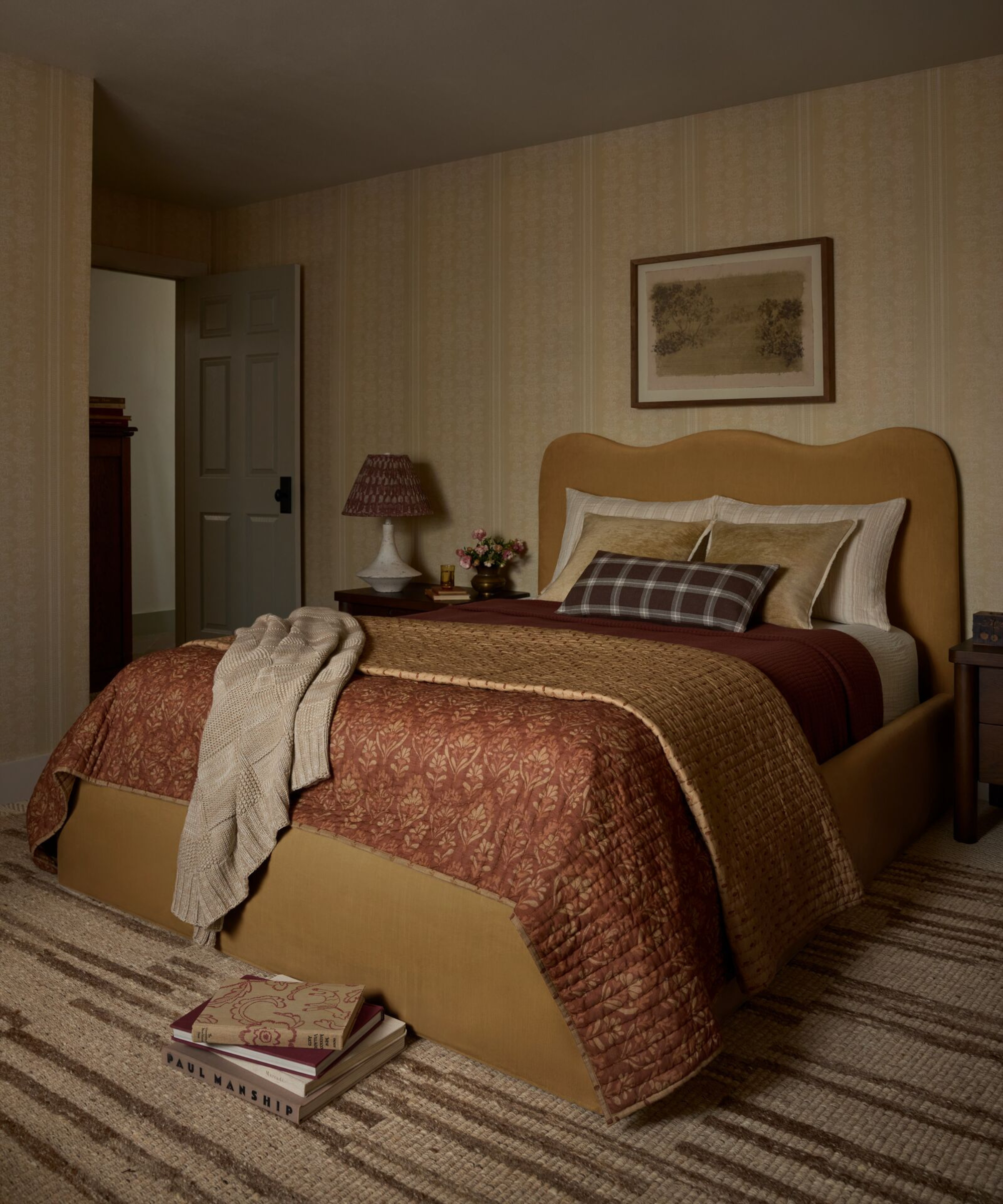 a warm toned bedroom with a red quilt folded at the foot of the bed, a mustard yellow upholstered bedframe, pillows, a striped rug on the floor and striped wallpaper on the walls