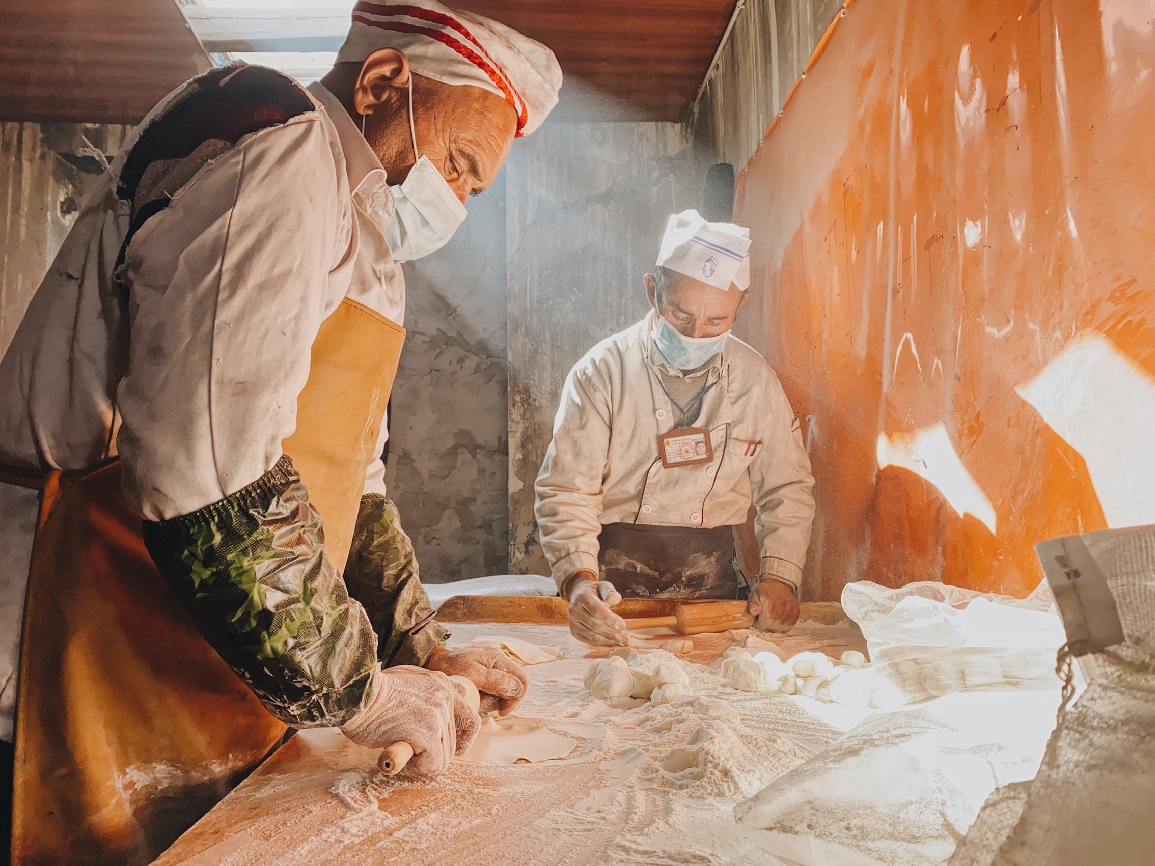 Two bakers in aprons and masks work diligently in a sunlit kitchen, rolling and shaping dough on a flour-dusted table