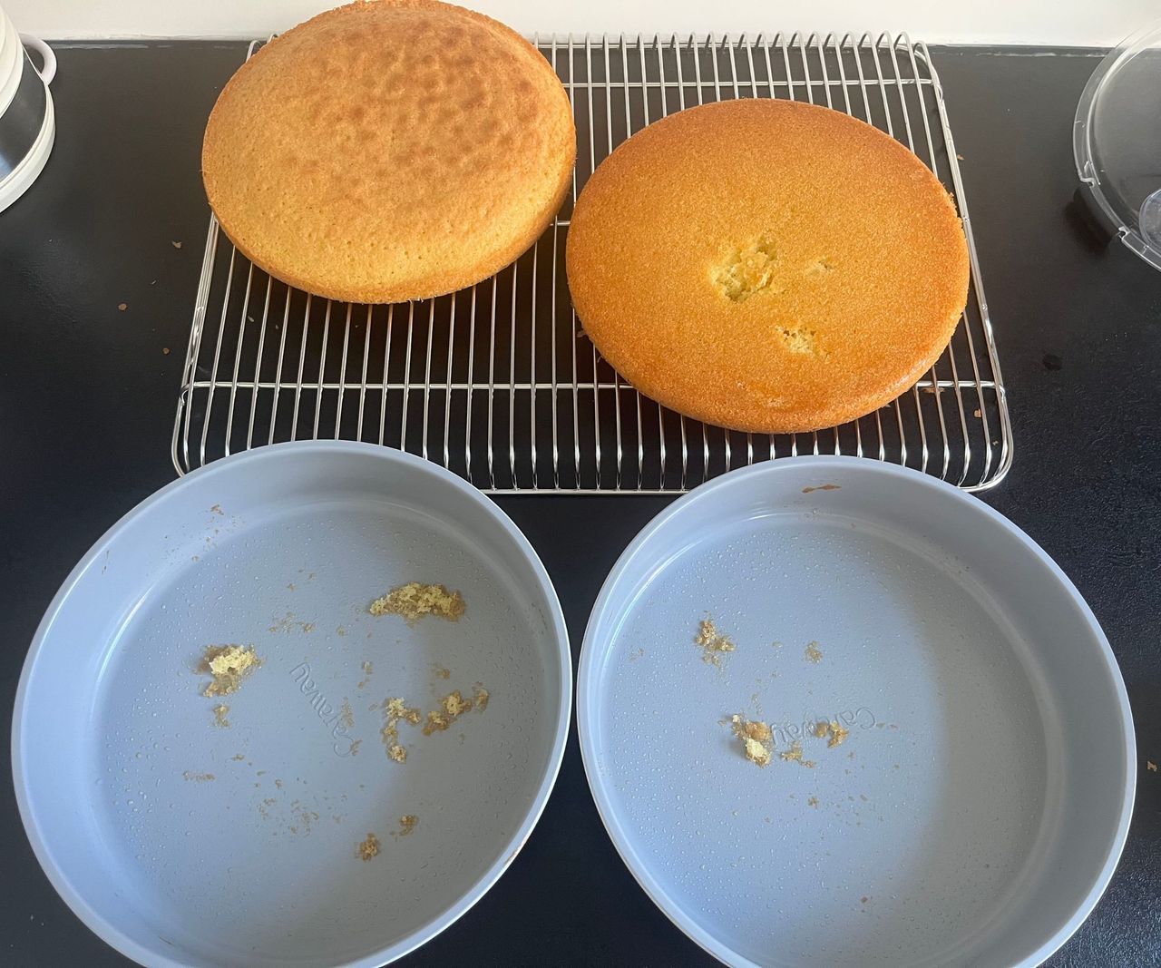Two golden sponges on a wire cooling rack with two grey cake tins in front of it