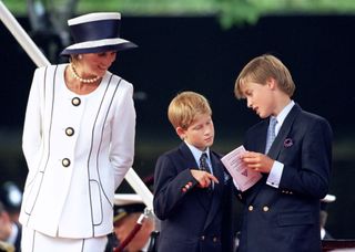 Princess Diana wearing a white skirt suit and navy hat smiling at Prince Harry and Prince William who are wearing navy suits and looking at a program