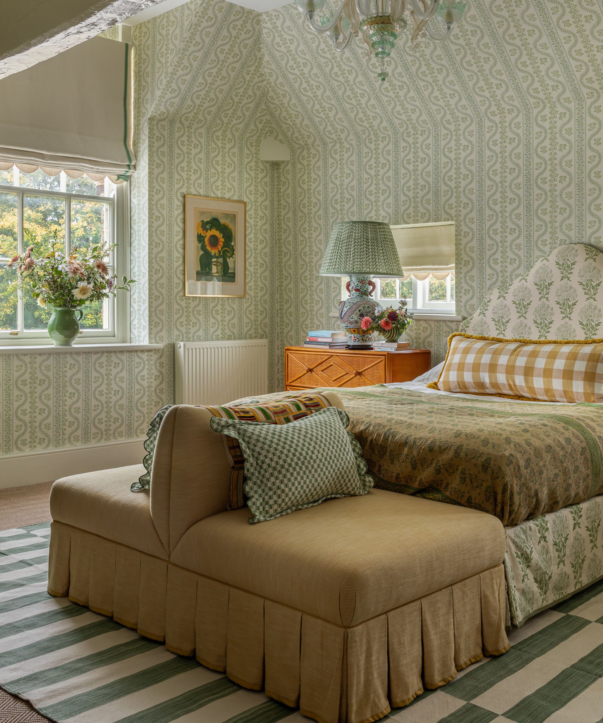 Bedroom with green and white wallpapered walls, an upholstered floral bed with a green quilt and yellow gingham cushion, a skirted yellow chaise lounge, a striped green rug, and a wooden nightstand.