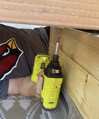 A person wearing t-shirt with Angry Birds motif lying on patio tiled floor using yellow electric drill to insert screw into outdoor sofa