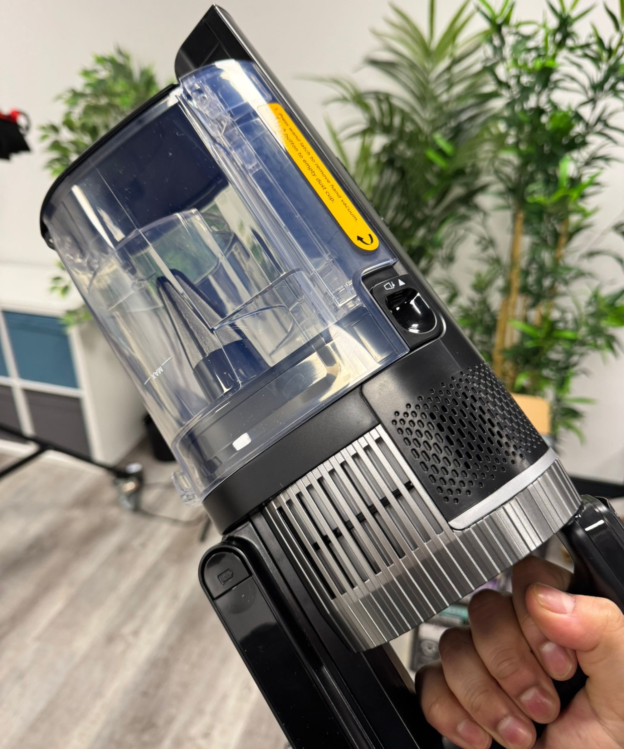 Hand holding the Shark Stratos Cordless Vacuum in front of tall green plants, white shelves and laminate flooring.