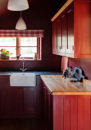 A traditional shaker style kitchen in red with a small window with a gingham blind above a butler sink.