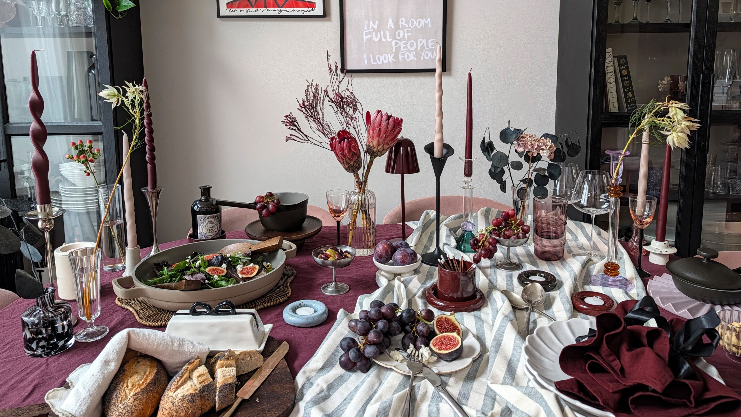 Table set for dinner party with burgundy and striped tablecloths, candlesticks, wireless lamp, vases of flowers, bowls of fruit