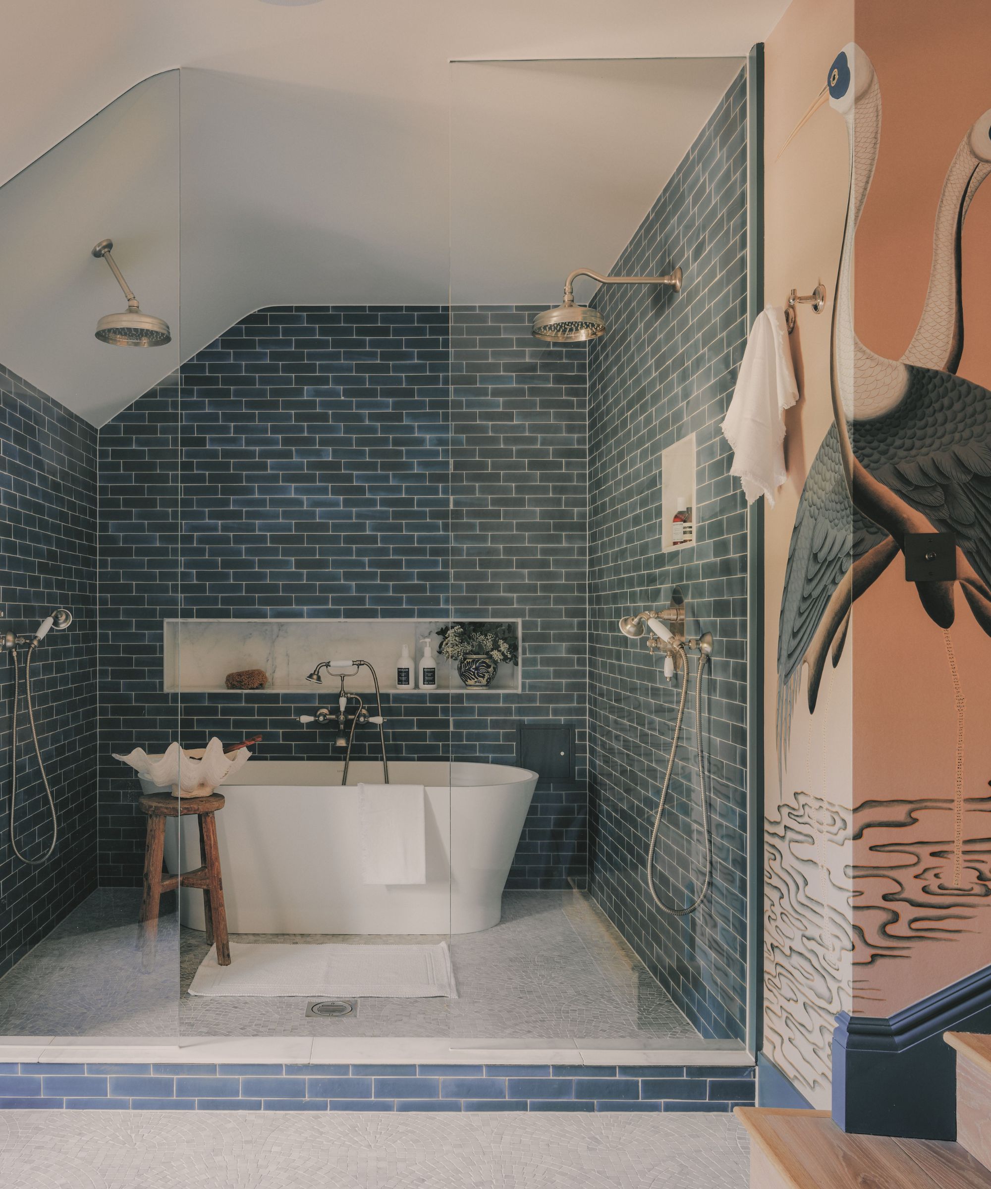 Walk-in shower room with dark blue subway tiles, dual shower heads, a freestanding white bathtub, and a wall featuring a large heron mural.