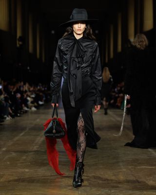 A model wearing a black hat, blouse, skirt, and tights walking at the Alexander McQueen F/W 25 show carrying a black handbag adorned with a red-and-black skull scarf.