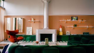 Image of a large living room with orange wallpaper and white paint in a color block, a green, glossy built-in, low cabinet, a green sofa, a chrome fireplace, and a red chair.