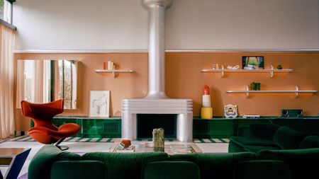 Image of a large living room with orange wallpaper and white paint in a color block, a green, glossy built-in, low cabinet, a green sofa, a chrome fireplace, and a red chair.