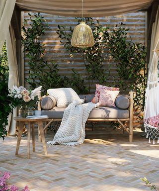 A backyard area with a gray bench with white and pink patterned throw pillows and a blanket, a side table with flowers on, with a pergola over it with beige curtains and a hanging rattan pendant light