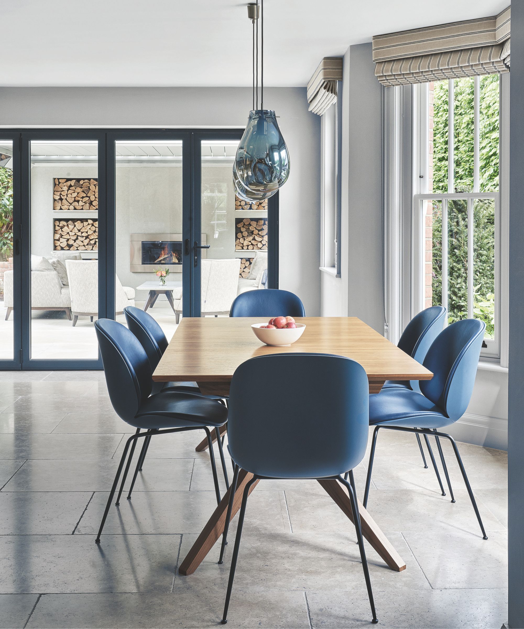A bright dining room with a wooden table and blue chairs, with a fruit bowl on top, tiled gray flooring, and a white fruit bowl on top of the table. Above the table are blue glass pendant lights, and to the right are bay windows with striped blinds. In the background are glass doors out onto a patio with a white wall, lit wood burner, log store, and white chairs.