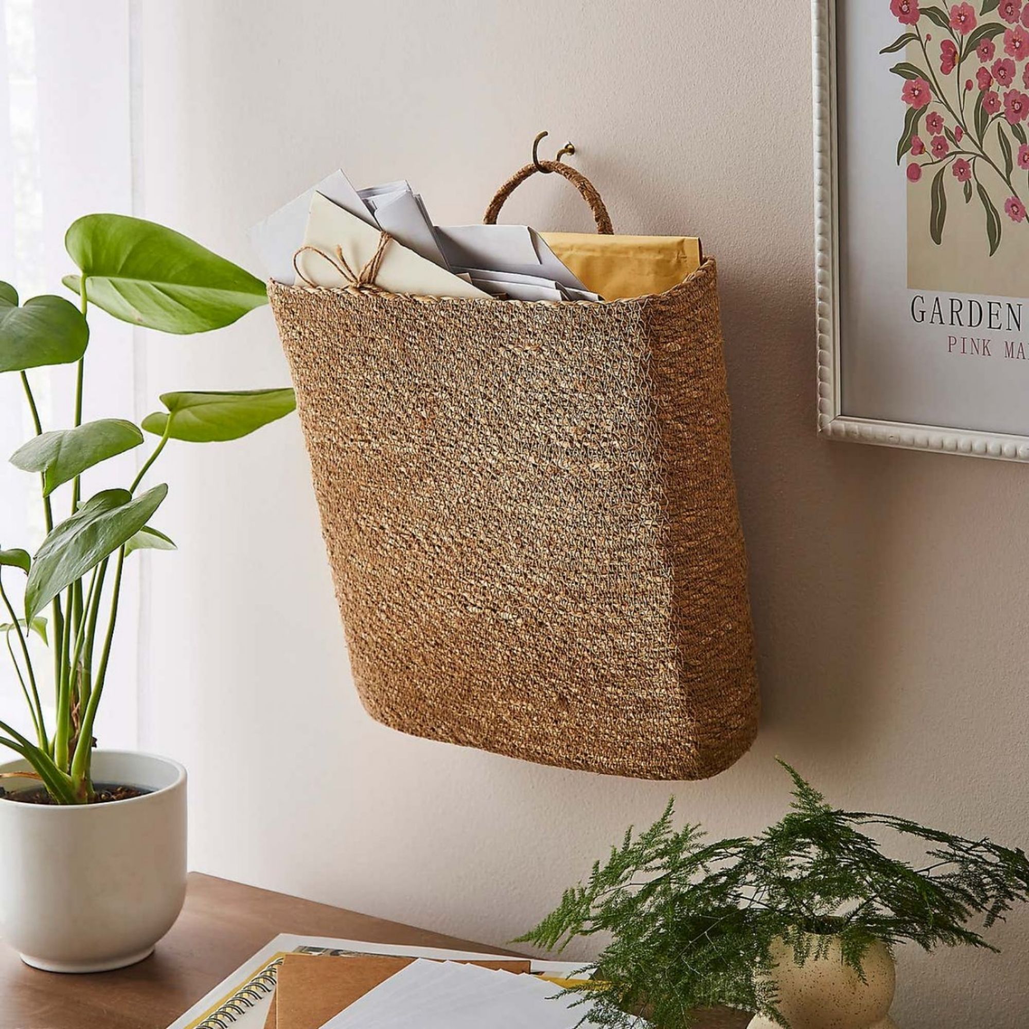 Dunelm Seagrass Letter Holder Basket hanging on a light pink painted wall, next to a photo frame and above a console table with plants on it