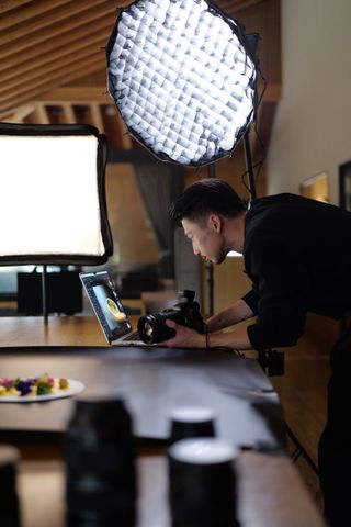 A person adjusting the LUMIX S1RII, while reviewing images on a laptop, set up in a well-lit studio with a table displaying food