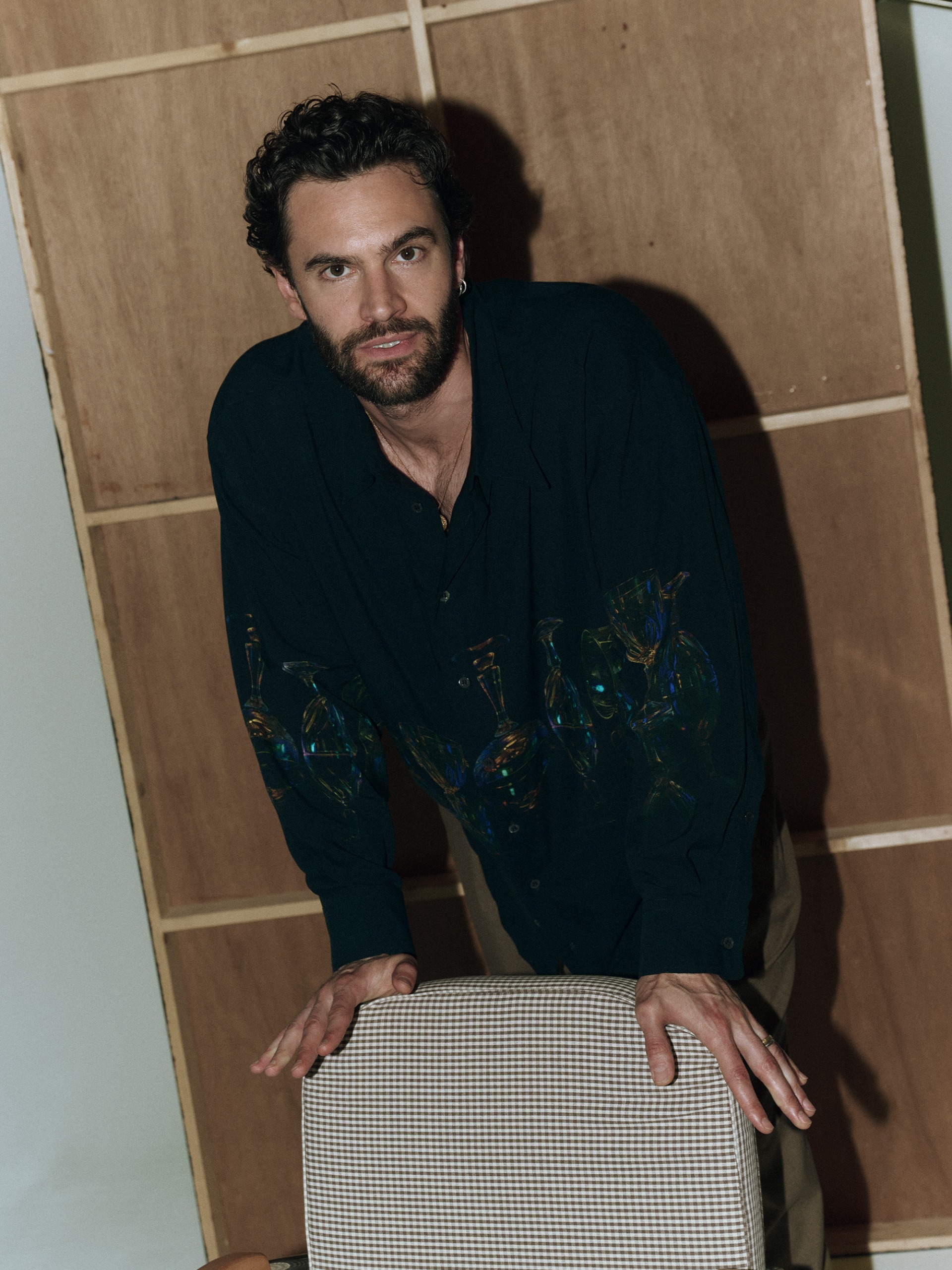 Tom Bateman photographed by Graham Dunn. He is leaning on a chair wearing a navy button-down shirt with different glassware printed on the front.