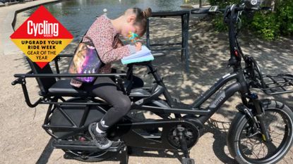 The Tern GSD electric cargo bike is side on with a passenger in the back seat who is writing in a pad. In the background there is gravel and a pond 
