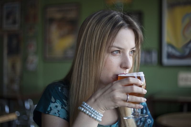 A blue-eyed woman sits, drinking a beer.