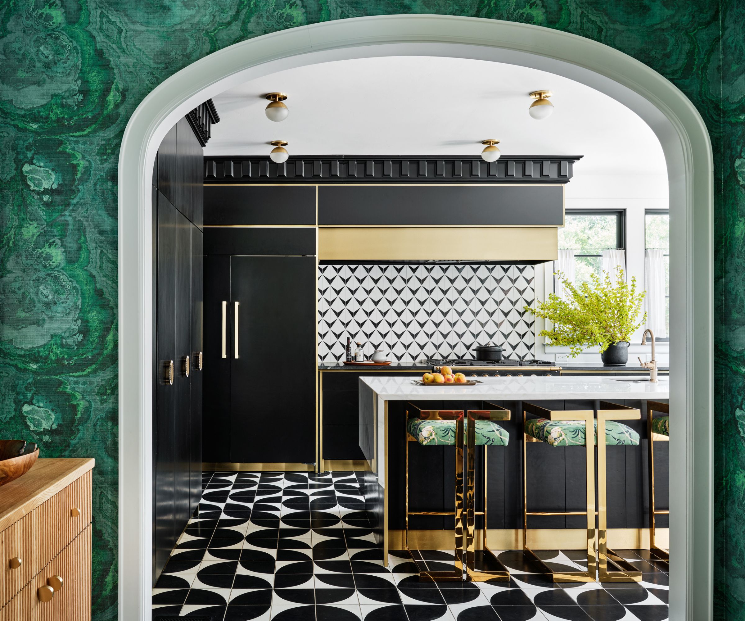 Arched doorway looking through into a modern kitchen with geometric tiled floor, black cabinets, a marble island with gold stools, and a tiled backsplash in black and white