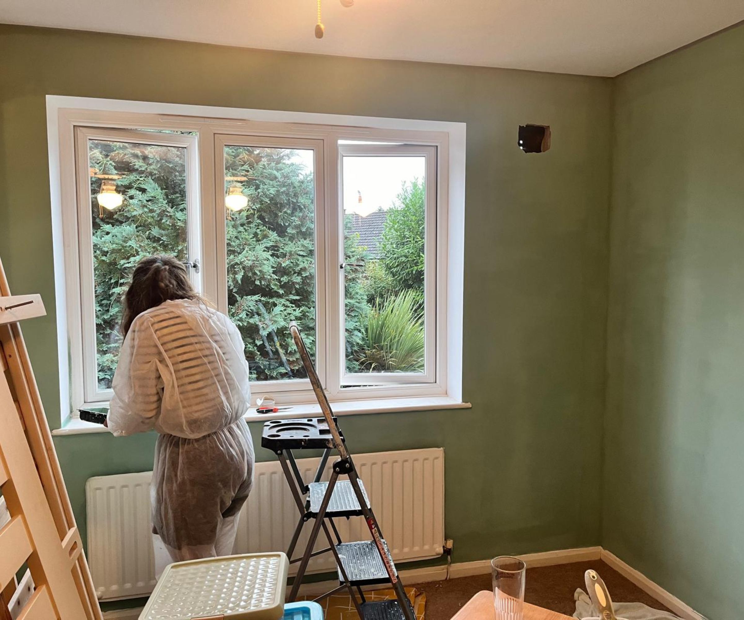 A mid-renovation bedroom, walls freshly painted sage green, with a person in a decorating suit painting the window frames on a stepladder.