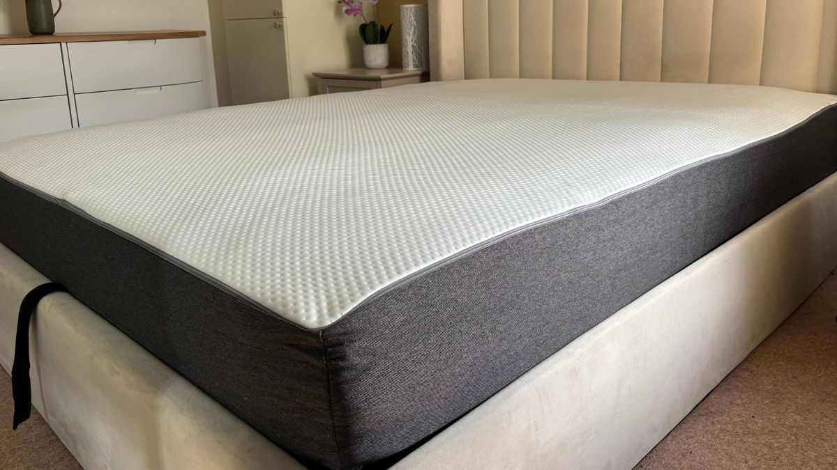 The image shows a grey and white mattress that is still expanding after being unboxed. It is placed on a grey fabric bed frame, set within a neutrally decorated bedroom