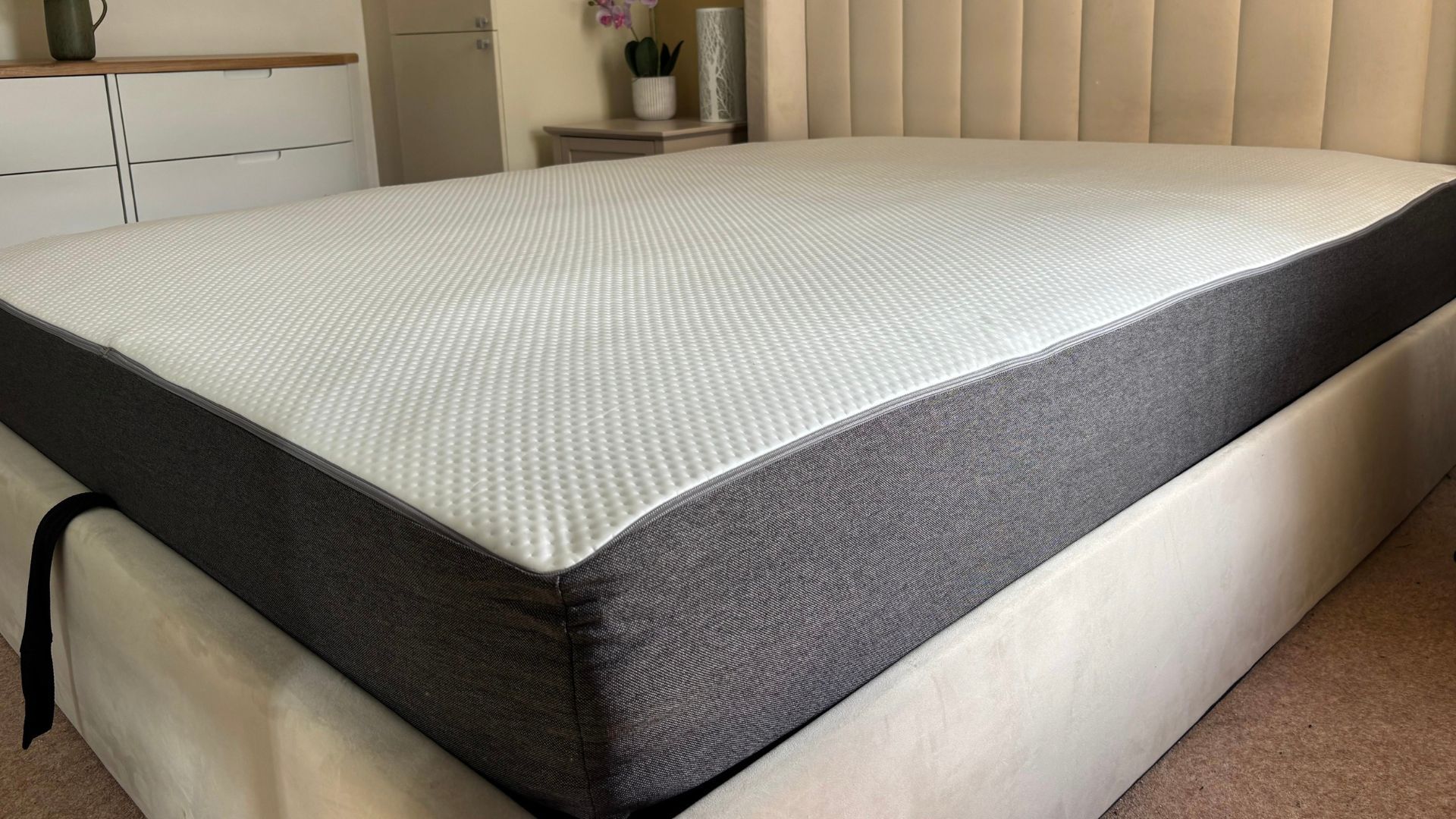 The image shows a grey and white mattress that is still expanding after being unboxed. It is placed on a grey fabric bed frame, set within a neutrally decorated bedroom